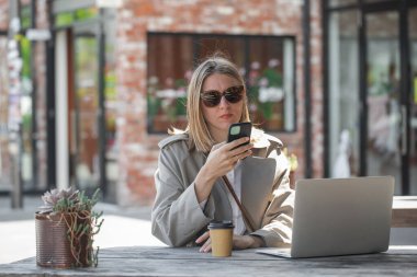 Kafe verandasında oturan telefon ve dizüstü bilgisayarla çalışan genç bir kadın. Uzak çalışma konsepti, iş kadını