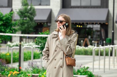Şehir merkezinde trençkotlu bir kadın telefonla konuşuyor. Şık bir kadın.