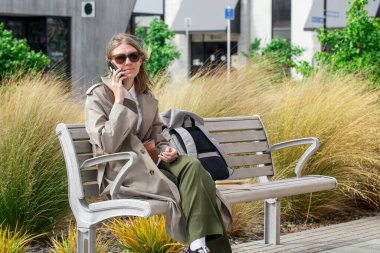 Trençkotlu kadın akıllı telefon ve sırt çantasıyla bankta dinleniyor. Yaşam tarzı.