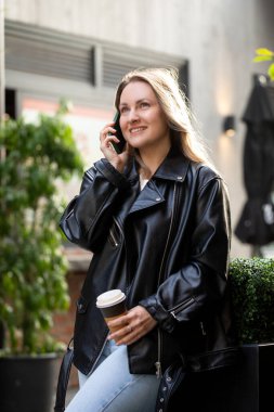 Deri ceketli beyaz bir kadın dışarıda telefonda bir fincan kahveyle konuşuyor. İş kadını.