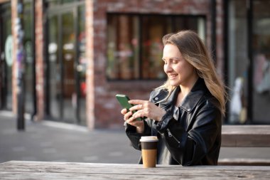 Kafenin açık verandasında bir fincan kahveyle otururken gülümseyen ve cep telefonuyla mesajlaşan genç güzel bir kadın. Genç beyaz kadın şehir ortamında kahve ve akıllı telefondan keyif alıyor.. 