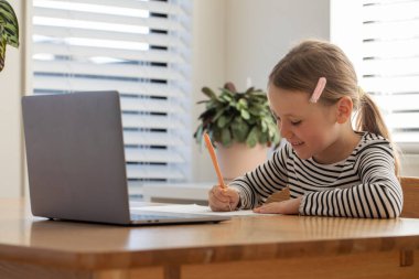 Evde dizüstü bilgisayar ve defterle e-öğrenim yapan mutlu genç bir kız. Eğitim kavramı.