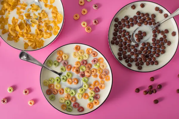 Farklı türde kahvaltı gevreği arka planında pembe arka planda süt var. Çikolata topları, mısır gevrekleri ve renkli yüzükler.