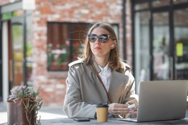 Moda bir kafede, şık bir kadın laptopuyla çalışıyor, lezzetli bir kahvenin ve açık hava ortamının tadını çıkarıyor.