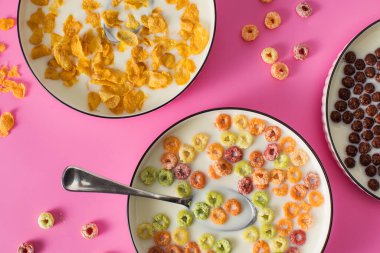 Farklı türde kahvaltı gevreği arka planında pembe arka planda süt var. Çikolata topları, mısır gevrekleri ve renkli yüzükler.