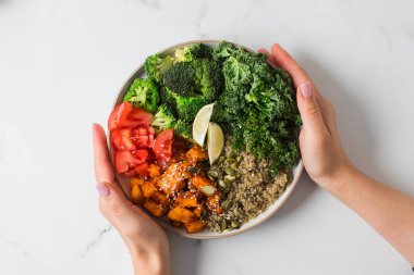 Bir kadının elleri mermer bir yüzeyde taze sebze ve kinoa dolu vegan bir Buda kasesini tutar.