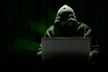 Kapüşonlu bir bilgisayar korsanı dizüstü bilgisayarla matris bilgisayar kodunun arka planına saldırıyor. Dijital çağda bilgisayar suçları kavramı.