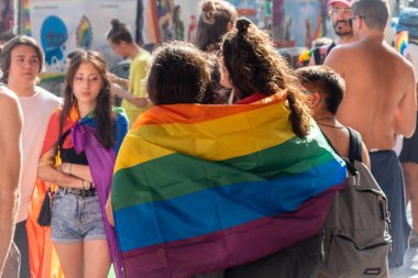 Barselona, İspanya - 15 Temmuz 2023: LGBTQ onur etkinliğinde düzenlenen protesto yürüyüşü sırasında sarılıp gökkuşağına sarmalanmış iki kişinin arka görüntüsü. Sahne çeşitliliği ve şenlik ruhunu gösteriyor.