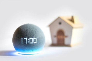 Alexa Echo Dot aygıtı mavi bir LED ışık etkinleştirilmiş ve arka planda bulanık bir ev figürü ile zamanı gösteriyor, akıllı bir evi simgeliyor..