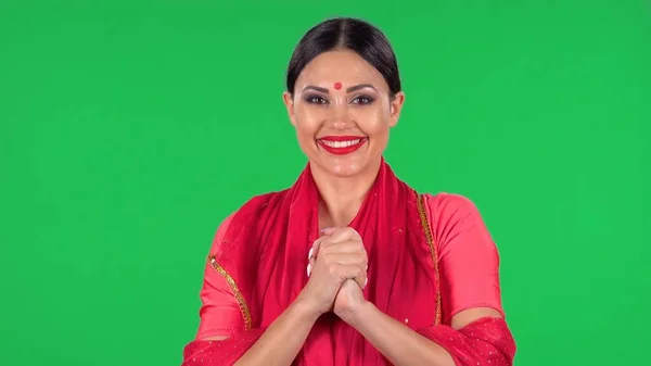 一个年轻的印度女孩的画像 红色的斑点缀在国家经典的红色莎丽直视 微笑和折叠的手掌到手掌 工作室的绿色荧幕上有一头长发和红唇的黑发 — 图库照片
