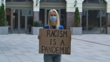 Tıbbi maske takan beyaz kadın protestocu, üzerinde RACISM bir PANDEMIC sloganı olan karton bir poster tutuyor. Karantina sırasında ırkçılığa ve şiddete karşı bir protesto. Irkçılık kavramını durdur