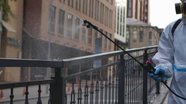 Beyaz koruyucu takım elbiseli ve solunum cihazlı bir sağlık çalışanı şehrin korkuluklarını dezenfekte ediyor. Virolog, alkol dezenfektanı ve fıskiyeyle antiseptik ile nesneleri tedavi ediyor. Coronavirüs