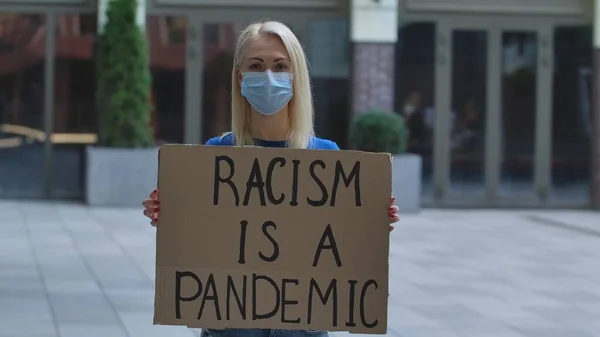 医療用マスクを着用した白人女性抗議活動家が Racism Pandemic というスローガンの段ボールポスターを持っている 隔離中の人種差別と暴力に対する抗議 人種主義の概念を — ストック写真