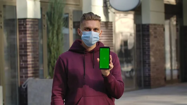 Portret Młodego Mężczyzny Masce Medycznej Wskazuje Smartfona Zielonym Kluczem Chromowym — Zdjęcie stockowe