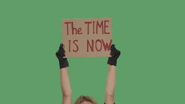 THE TIME IS NOW. Protest text message on cardboard. Stop racism. Police violence. Banner design concept. Hands in black gloves holding a poster a green screen, chroma key