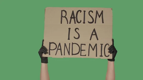 Showing cardboard poster with inscription RACISM IS A PANDEMIC. Human Rights Demonstration. Hands in black gloves holding a poster on green screen, chroma key. Stop Racism concept, No Racism