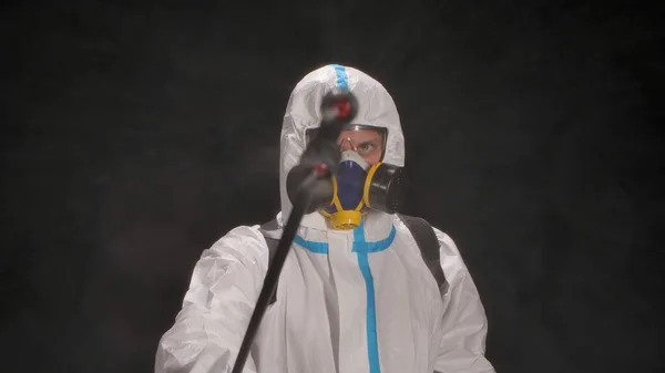 Man in a protective suit with a high-pressure apparatus disinfects with an antiseptic spray. Medical worker sprays disinfectant spray while looking at camera on black background. Concept of sanitation