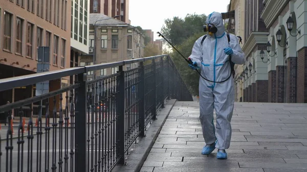 Medical worker in white protective suit and respirator disinfects railing on city street. Virologist treating objects with an antiseptic using an alcohol disinfectant and sprinkler. Coronavirus