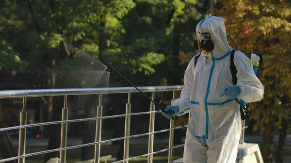 A virologist in a white protective suit and respirator disinfects railings on city streets. Disinfection of objects with antiseptic spray to prevent the spread of the Covid 19 outbreak