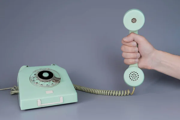 男性の手は灰色の背景にロータリー電話の近くに古い青のプラスチック製の電話受信機を保持 男の手の中にレトロな家庭用電話装置からのリモート携帯電話を閉じます コミュニケーションの概念 — ストック写真