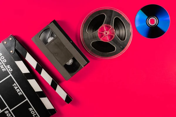 Compact disc, filmstrip bobbin, black plastic VHS video cassette and clapper board on pink background. Old storage media, drives for watching videos or movies or listening to music. Analogue film and