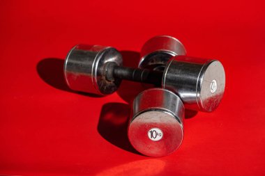 Kırmızı arka planda uzanan iki gümüş halter ve gölgeler. Dambılların her biri kasları şişirmek için on kilogram ağırlığında. Spor aletleri, vücut geliştirme, yaya geçidi. Konsept