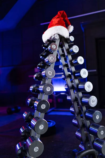 クリスマスツリーと赤いサンタクロースの帽子の形で金属ダンベルのセットが付いているラック 青い光の中でジムでのスポーツ機器 クロスフィット ボディビルディング フィットネス用のダンベル バーベル — ストック写真