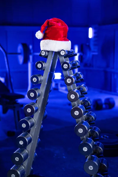 クリスマスツリーと赤いサンタクロースの帽子の形で金属ダンベルのセットが付いているラック 青い光の中でジムでのスポーツ機器 クロスフィット ボディビルディング フィットネス用のダンベル バーベル スポーツベンチ — ストック写真