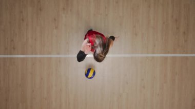 Genç bir kadının ahşap zeminde topa vurup zıplayarak topa vuruşunun en üst görüntüsü. Bayan sporcu, yarışmadan önce top servis etme becerilerini geliştirir. Sporcu kadın voleybol antrenmanında.