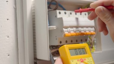 Voltaj altında çalışan elektrikçi, elektrikçilerin kullanılabilirliğini kontrol ederek voltajı sarı multimetreyle ölçüyor. Bir adamın elleri beyaz düğme kutusunu test ederken voltaj santralini kapatır.