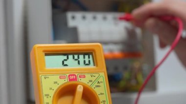 Voltaj altında çalışan elektrikçi, elektrikçilerin kullanılabilirliğini kontrol ederek voltajı sarı multimetreyle ölçüyor. Bir adamın elleri beyaz düğme kutusunu test ederken voltaj santralini kapatır.