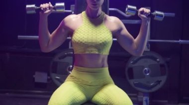 Sarı spor kıyafetli vücut geliştirici kadın dambılları kaldırıyor ve nefes alıyor. Antrenman omuzları. Spor salonunda mavi neon ışığıyla kas egzersizi yapan kadın sporcu. Güzel, parlak bir vücut. Kapat.