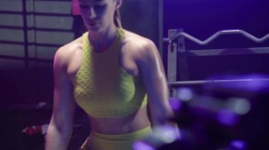 Sarı spor kıyafetli vücut geliştirici kadın dambılları kaldırıyor ve nefes alıyor. Antrenman omuzları. Spor salonunda mavi neon ışığıyla kas egzersizi yapan kadın sporcu. Güzel, parlak bir vücut. Kapat.