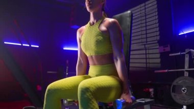 Sarı spor giysili spor elbiseli kadın spor bankında oturur, dambıl alır, omuzlar için egzersiz yapar, nefes alır. Spor salonundaki kadın sporcular arka planda halterli, ağırlıklı ve...