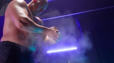 Çıplak gövdeli kaslı bir adam beyaz tozla ellerini çırpıyor ve ağır halterleri siyah ağırlıklarla kaldırıyor. Vücut geliştirici engelleri itiyor ve sırt egzersizleri yapıyor. Güç kaldırma ya da geçiş