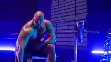 Çıplak gövdeli vücut geliştirici eliyle ağır dambıl kaldırıyor ve pazılar ve trisepsler için egzersiz yapıyor. Spor salonundaki spor bankında oturan kaslı adam arka planda, halterli ve...