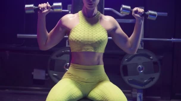 身穿黄色运动服的健美女子举起哑铃和呼吸 训练的肩膀女运动员在健身房用蓝色霓虹灯进行肌肉锻炼 美丽闪亮的身体皮肤 靠近点 — 图库视频影像