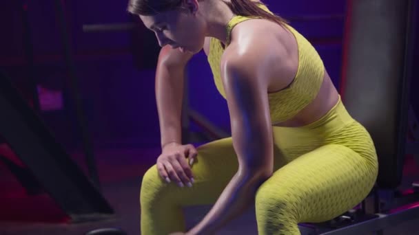 健康的女人穿着黄色运动服 用哑铃训练 做二头肌运动和呼吸 女运动员坐在体育馆的运动长椅上 身上点着蓝色霓虹灯 美丽闪亮的肌肤 — 图库视频影像