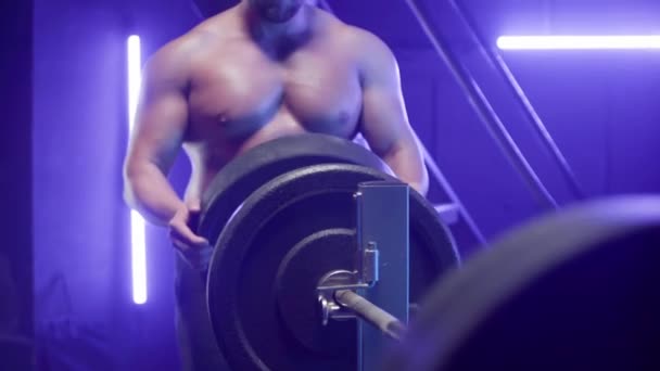 肌肉发达的男性健美运动员 赤身裸体的躯干在杠铃上缠绕着黑色的体重 体操比赛中肌肉鼓鼓的男人 蓝光准备力量训练 动力提升的概念 — 图库视频影像