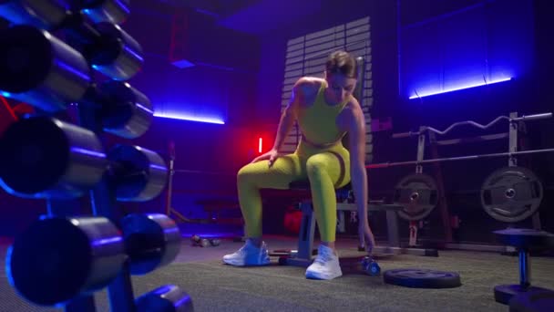 健康的女人穿着黄色运动服坐在运动长椅上 拿哑铃 做肩膀运动 女田径运动员在体育馆里 背靠着哑铃 举重和滑翔伞进行训练 — 图库视频影像