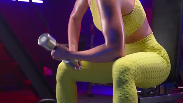 穿着黄色运动服 带哑铃 做二头肌运动的健康女性 女运动员坐在体育馆的运动长椅上 身上点着蓝色霓虹灯 美丽闪亮的身体皮肤 躯干靠拢 — 图库视频影像