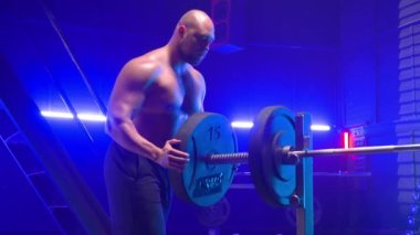 Çıplak gövdeli kaslı erkek vücut geliştirici siyah ağırlıkları halterin üzerine diziyor. Spor salonunda kasları şişmiş bir adam mavi ışık ile güç antrenmanına hazırlanıyor. Spor anlayışı, güç kaldırma