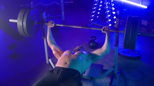 健美运动员躺在运动长椅上 推着重磅的杠铃 然后把它放回架子上 肌肉男在带哑铃 杠铃和哑铃的机架的背景下从事举力运动 — 图库视频影像