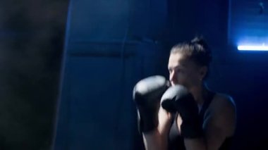 Kickboks antrenmanında mavi ışıkla. Siyah üniformalı, boks eldivenli ve bacak korumalı genç bir kadın boksör kum torbasına güçlü yumruklar atıyor. Spor, eylem, güç, enerji