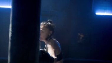 Kickboks antrenmanında mavi ışıkla. Siyah üniformalı, boks eldivenli ve bacak korumalı genç bir kadın boksör kum torbasına güçlü yumruklar atıyor. Spor, eylem, güç, enerji