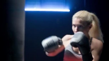 Kickboks, kadın boksör yumruklarını eğitir. Kırmızı spor üniformalı ve siyah boks eldivenli genç sporcu lacivert ışıklı kum torbasını dövüyor ve tekmeliyor. Kickbox antrenmanı. Kutu