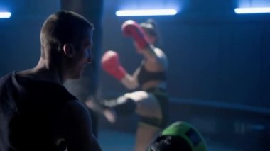 Sinematik kickboks çekimi. Ön planda kadın boksör antrenörle yumruk ve tekme atmayı öğretiyor. Arka planda, bir bayan atlet kum torbasına vurma alıştırması yapıyor. Boksta kickboksçular