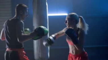 Kickboks eğitimi. Boks eldivenli kadın, koyu renk spor salonunda mavi ışıkla antrenörüyle boks yapıyor. Kadın kickboksör ve erkek çalıştırıcının antrenman maçı. Dişi dövüşçü yumruk ve tekme pratiği yapar.