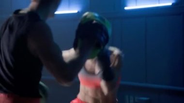 Kickboks eğitimi. Boks eldivenli kadın, koyu renk spor salonunda mavi ışıkla antrenörüyle boks yapıyor. Kadın kickboksör ve erkek çalıştırıcının antrenman maçı. Dişi dövüşçü yumruk ve tekme pratiği yapar.