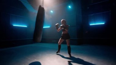 Kickboks antrenmanında mavi ışıkla. Siyah üniformalı, boks eldivenli ve bacak korumalı genç bir kadın boksör kum torbasına güçlü yumruklar atıyor. Spor, eylem, güç, enerji